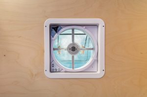 A Maxxair Maxxfan Deluxe fan installed in the ceiling of a camper van with a baltic birch wood ceiling