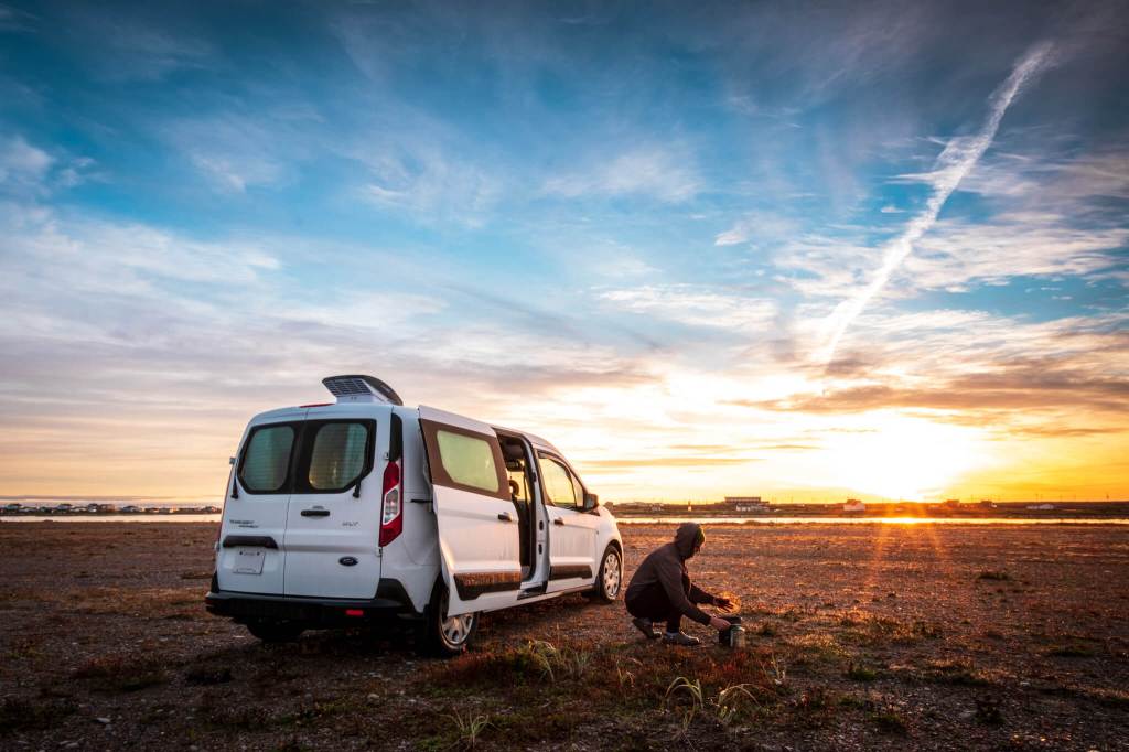 Person making coffee on a camp stove beside a Transit Connect camper van at sunrise in Flower’s Cove, Newfoundland
