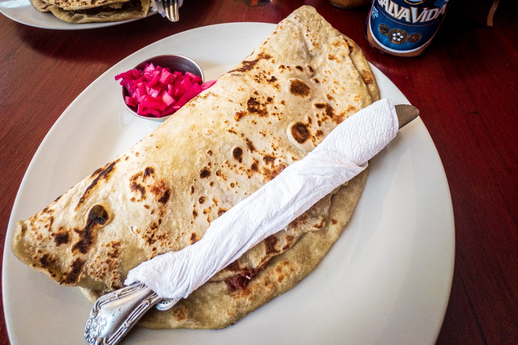 A baleada on a plate with utensils, pickled red onion and can of local beer