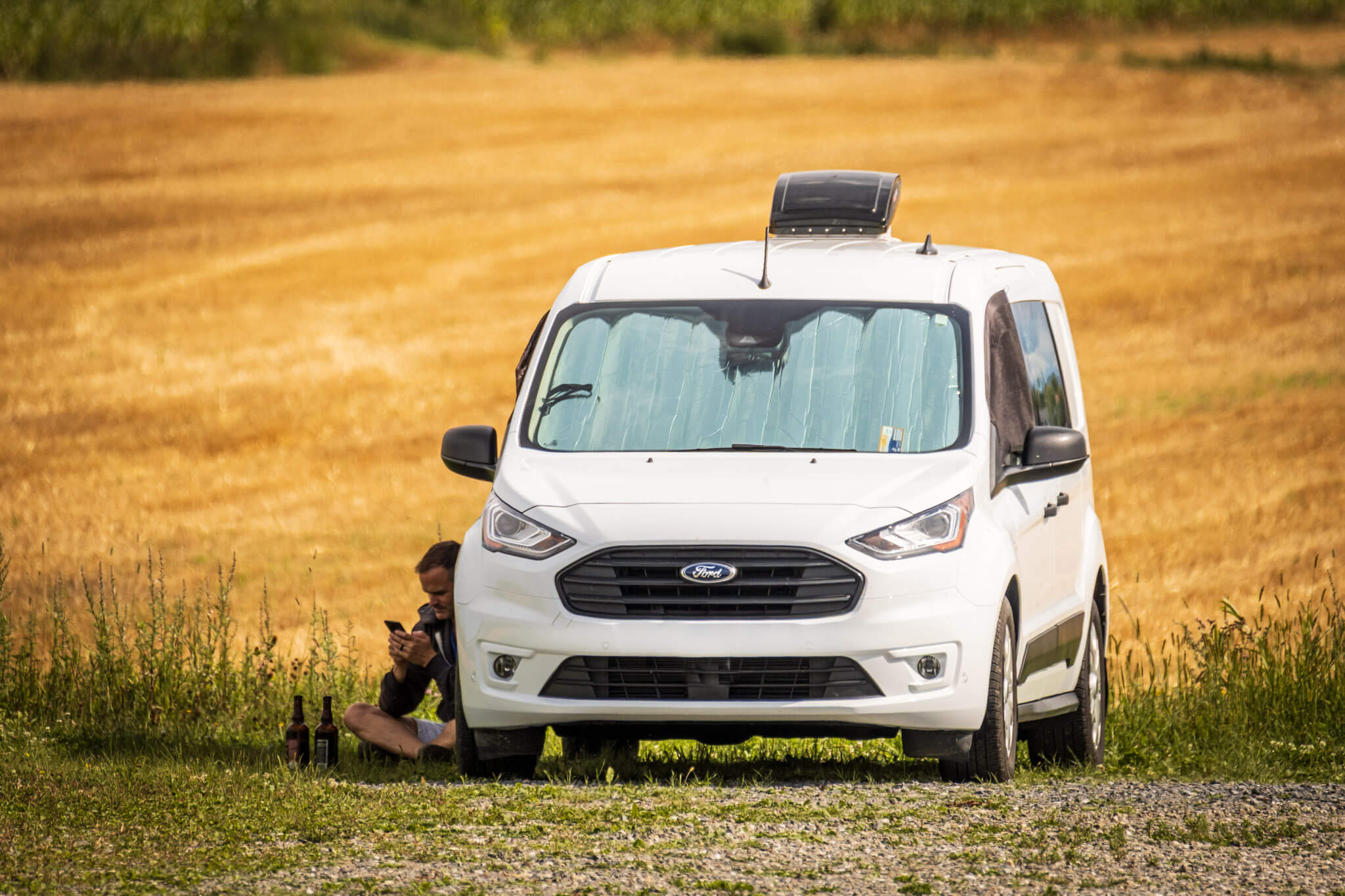 Camper sitting in the shade of a Ford Transit Connect camper van near a wheat field